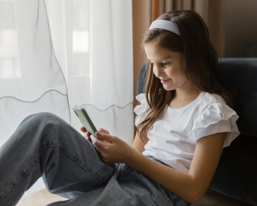 Teenage girl with long brown hair, sitting near the window and looking at her smartphone.